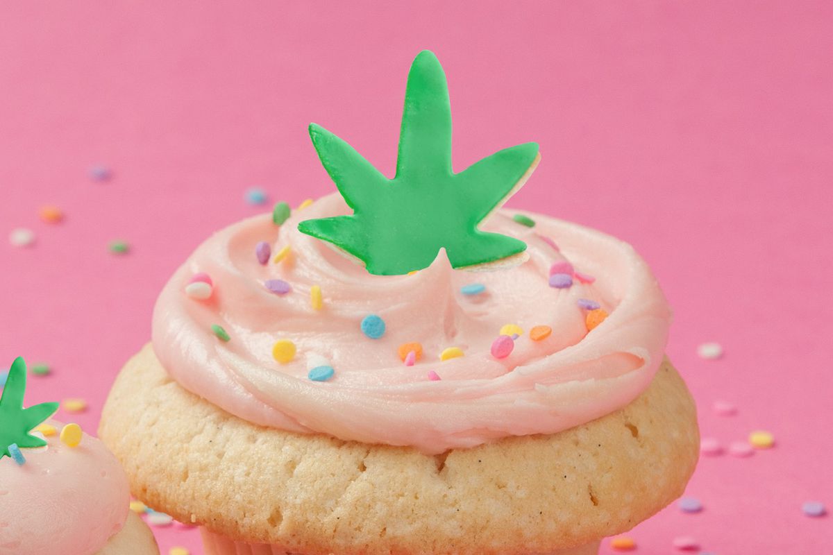A cupcake with a marijuana leaf decoration on top against a pink background 