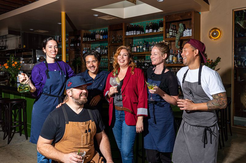 A racially and gender-diverse group wears aprons and smiles together inside the restaurant. 