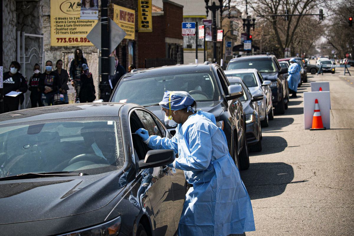 People sit in a line of cars while masked and gowned health care workers approach to test for the coronavirus.