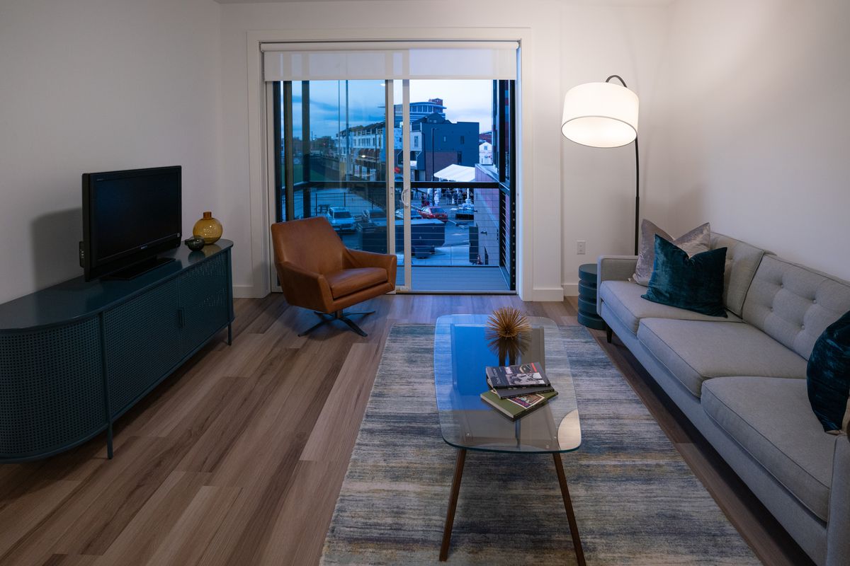 A small TV sits on a black stand across from a white couch. There’s a narrow glass coffee table on top of wood plank flooring. A sliding doors leads to a little balcony. 