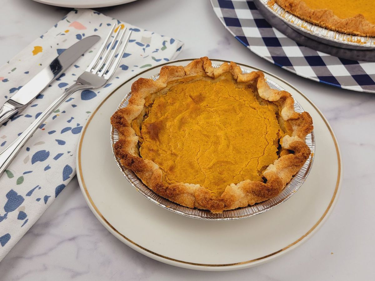 A whole pumpkin pie sitting on a white plate next to a fork and knife on a paint-splattered napkin