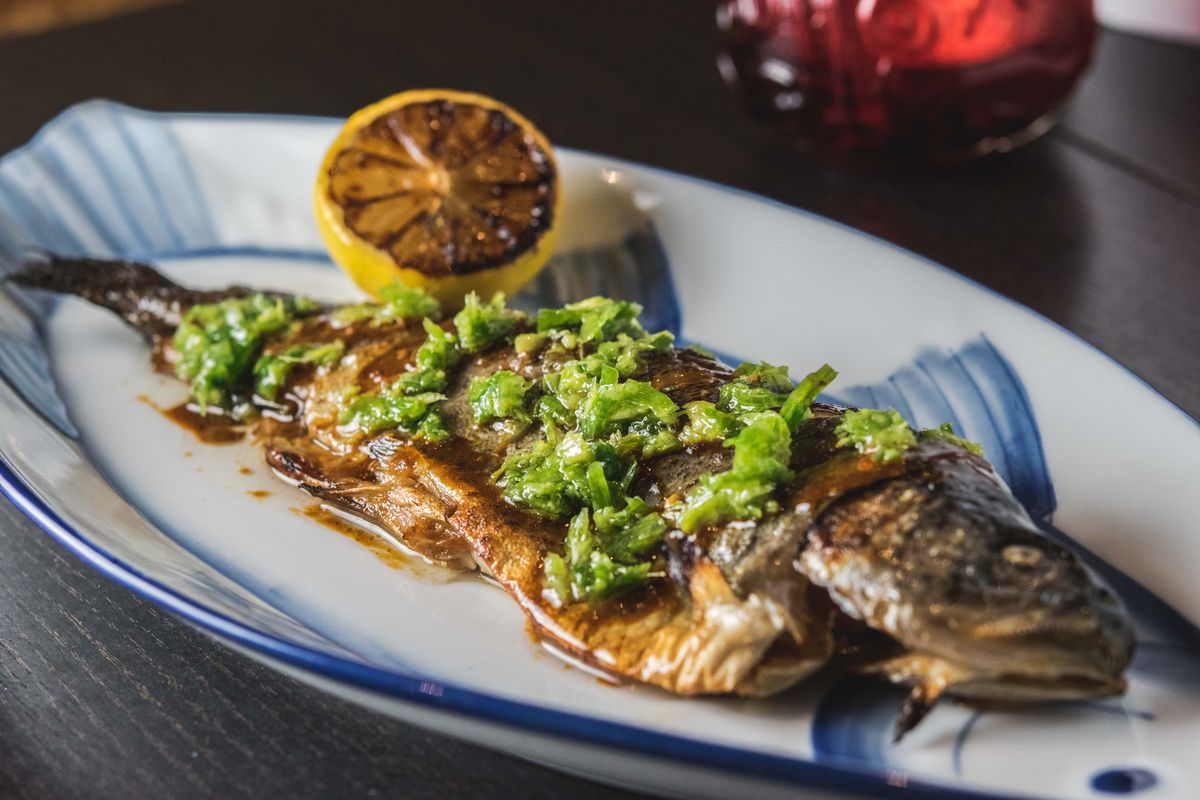 A whole trout sits on a plate, topped with a green sauce and accompanied by a half a grilled lemon