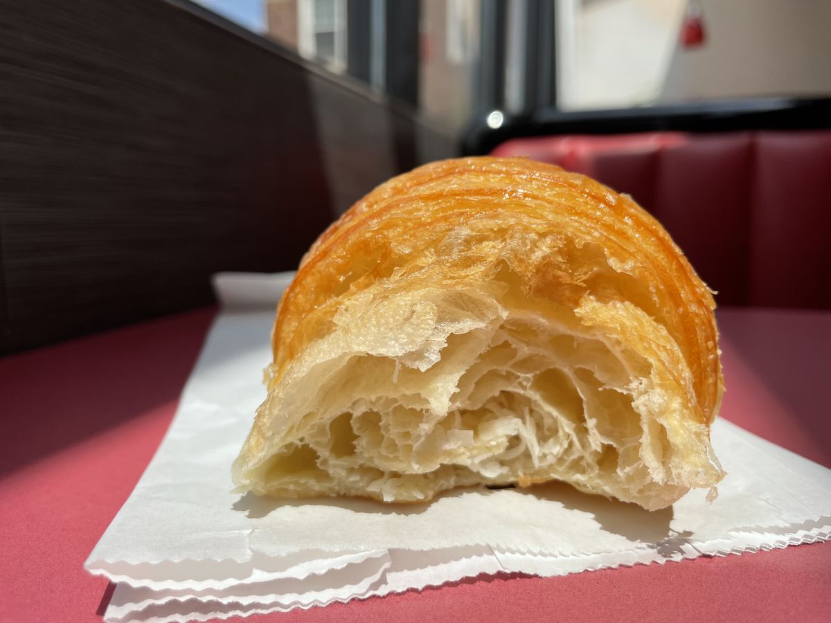 A flaky croissant that’s half bitten into sits on a white paper bag on a red table.