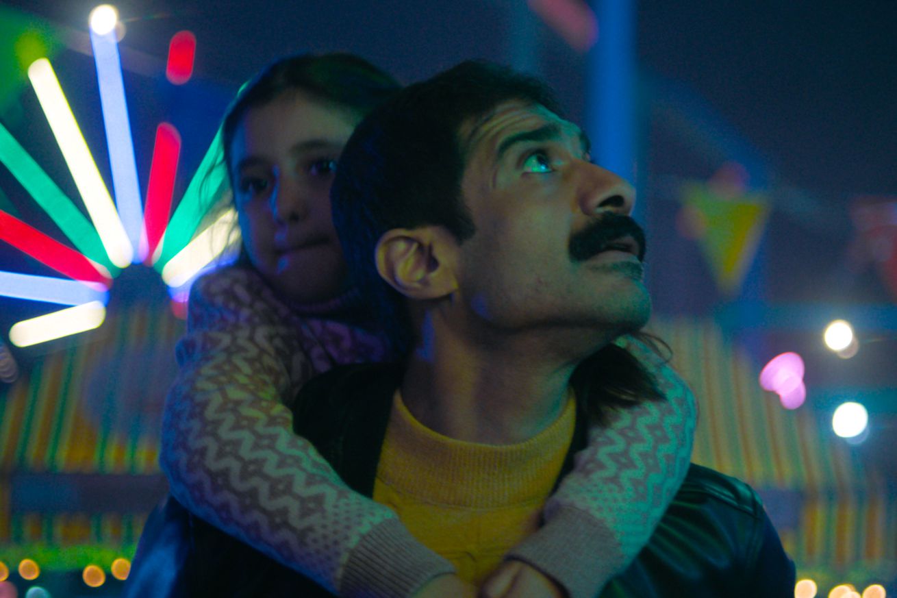 A mustachioed man wearing a yellow sweater and a black leather jacket. The man is carrying a young girl in a white sweater on his back, and the pair are standing together at a carnival.