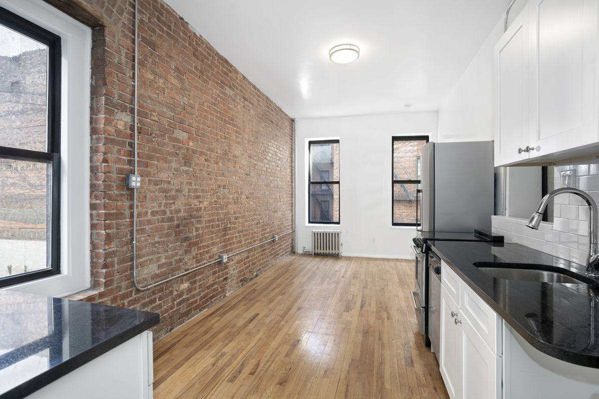 A living area with exposed brick, hardwood floors, and a kitchen with white cabinetry.