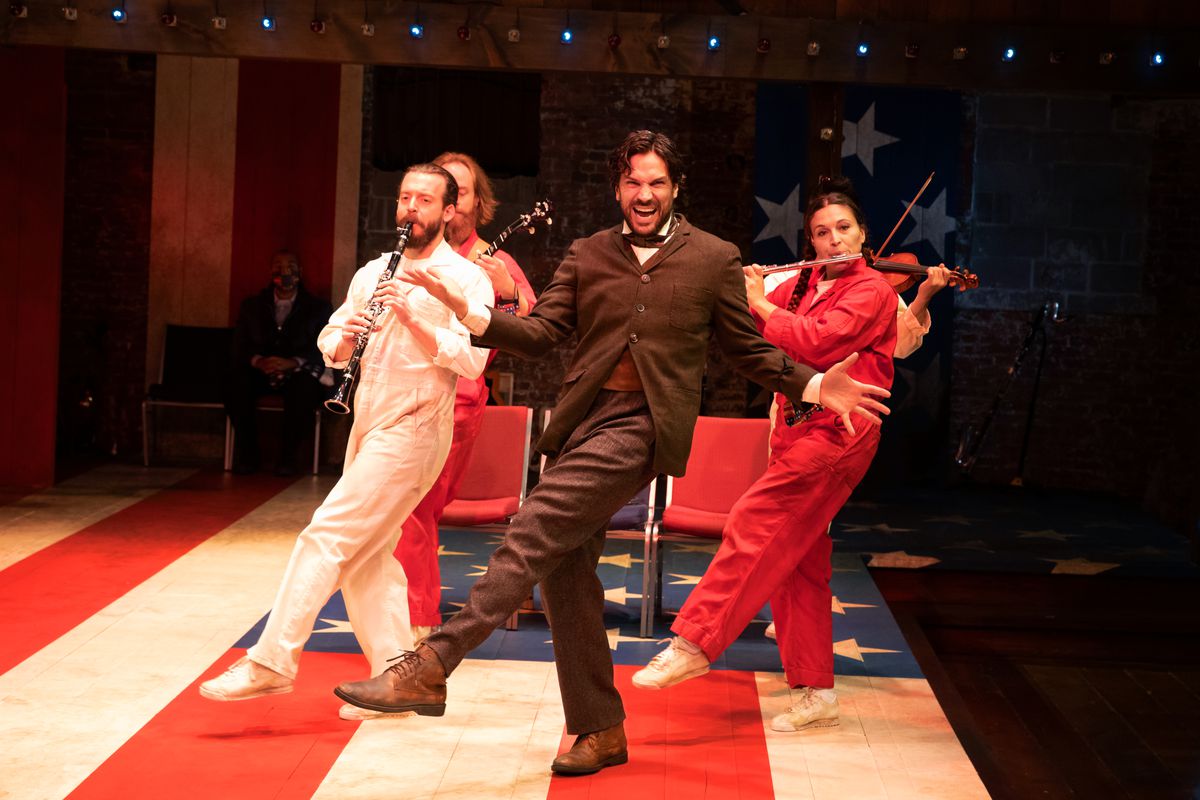 A man in a suit is dancing in front of a trio of musicians. The musicians are wearing prison-style jumpsuits in red, white, and blue. The dancing man in the suit is smiling hugely but his eyes are panicked.