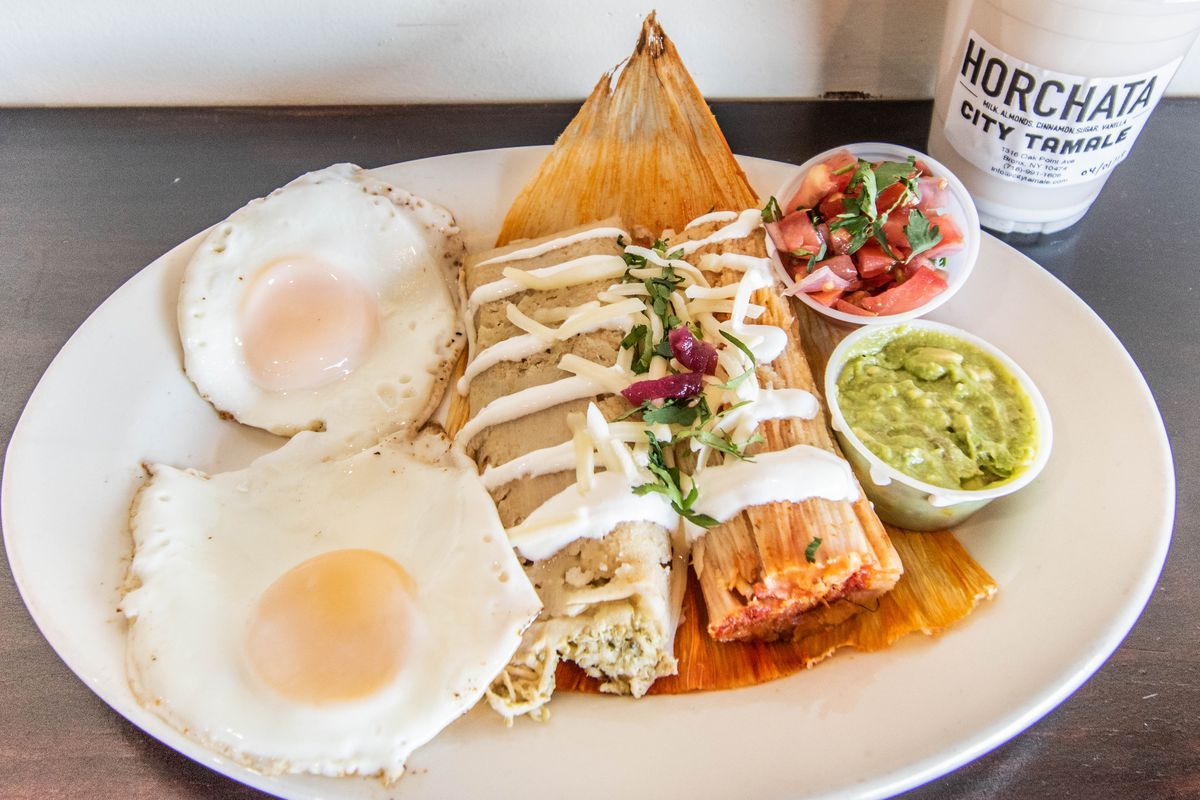 Two tamales are covered in crema and pico de gallo and accompanied by two fried eggs, a side of guacamole, and a side of salsa