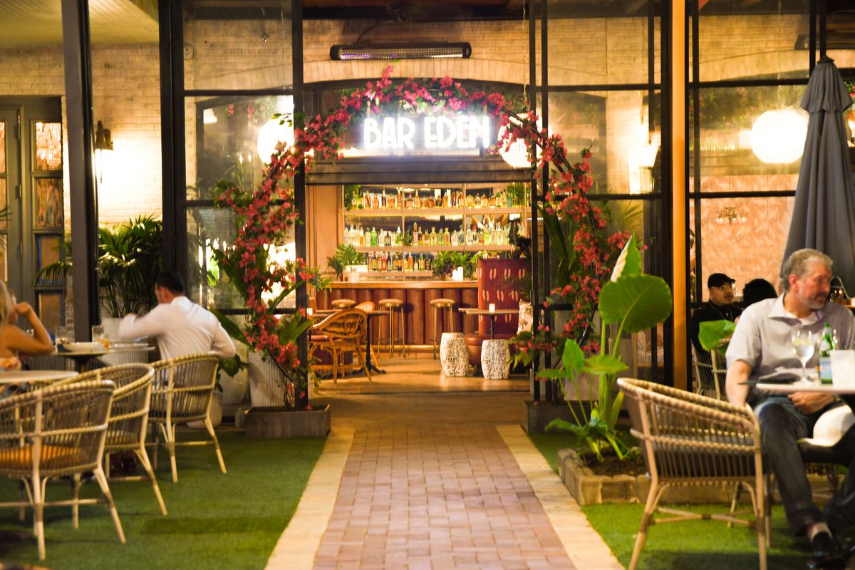 A bar exterior shows people sitting in the garden outside with cocktails, in wicker chairs. A floral arch with dark pink flowers is over the glass door with black frame. Inside the bar beckons and above it is a white neon sign that reads: Bar Eden.