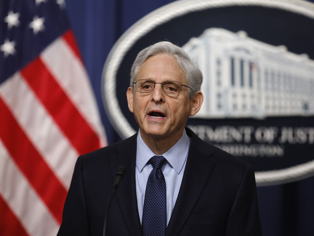 Attorney General Merrick Garland speaks in front of a large Department of Justice seal, flanked by a US flag.
