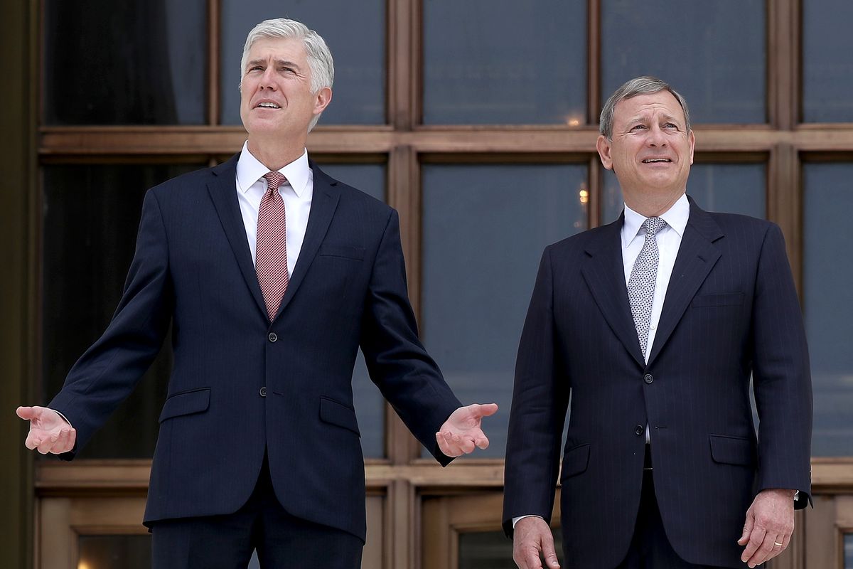 Gorsuch (left) raises both hands in a gesture. Both men wear black suits. They stand in front of a large window divided by wooden window frames.