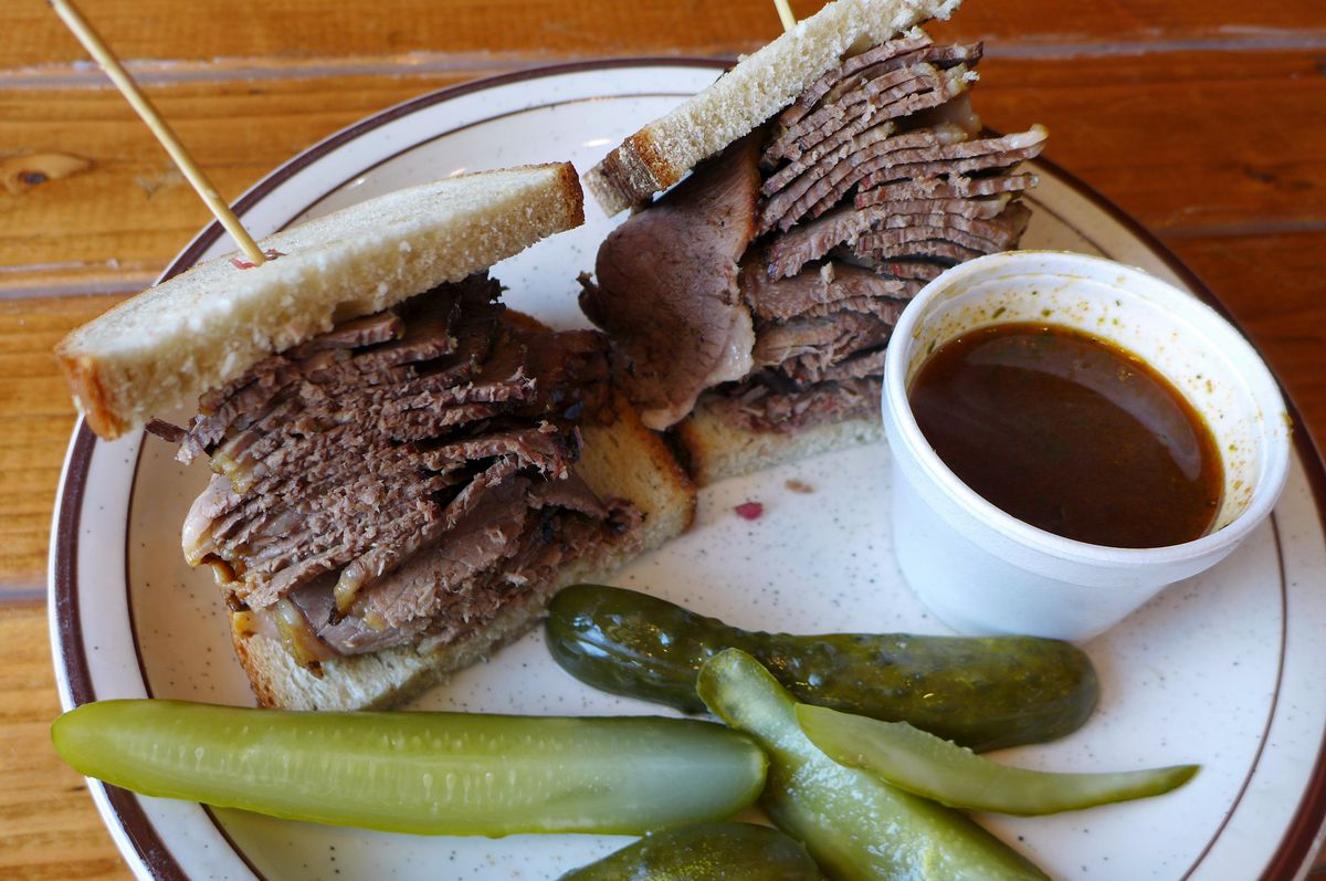 A meat-filled sandwich with pickles and a cup of gravy on the side.