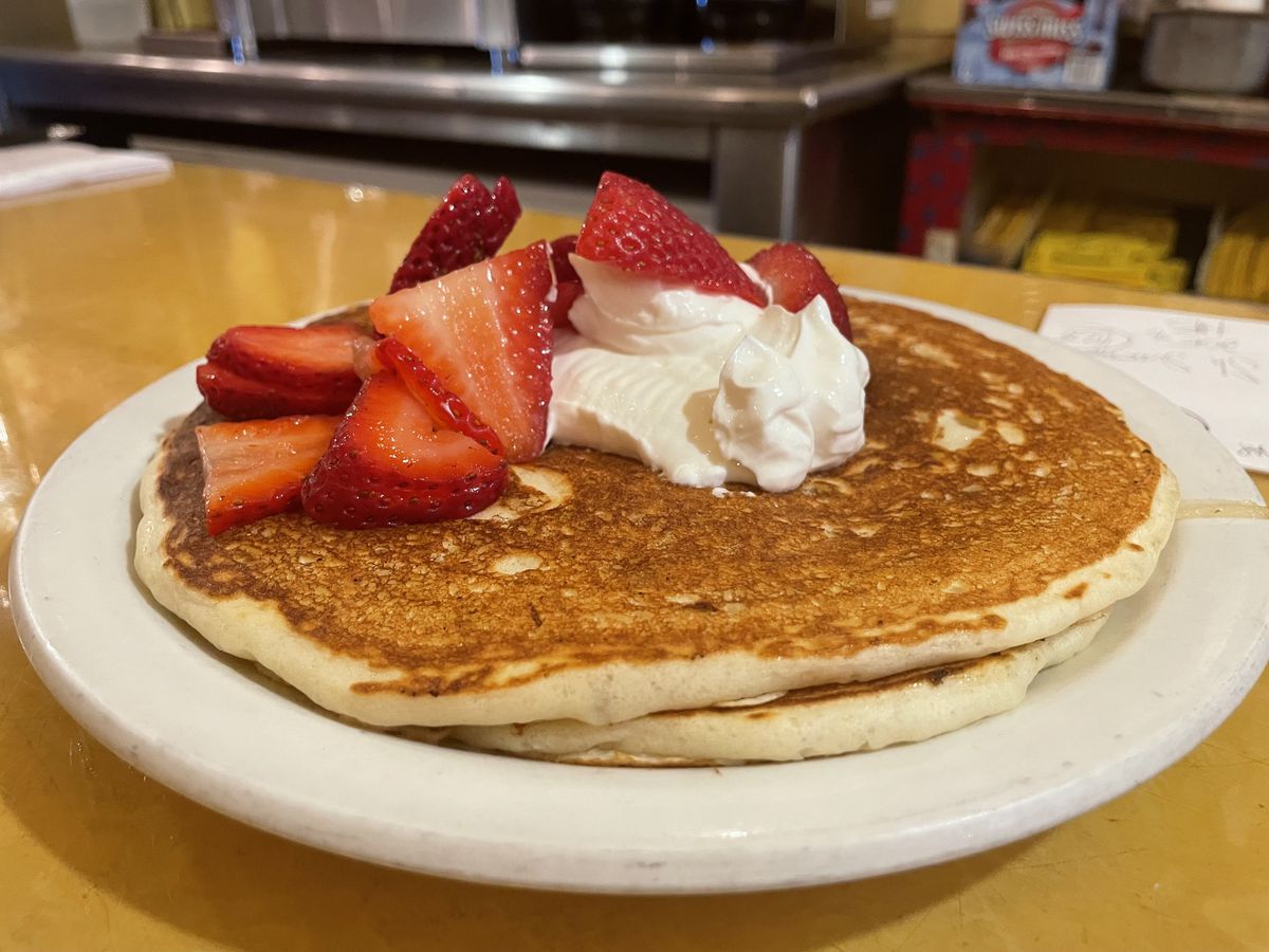 Two pancakes on a white plate topped with sour cream and strawberries on a yellow counter. 