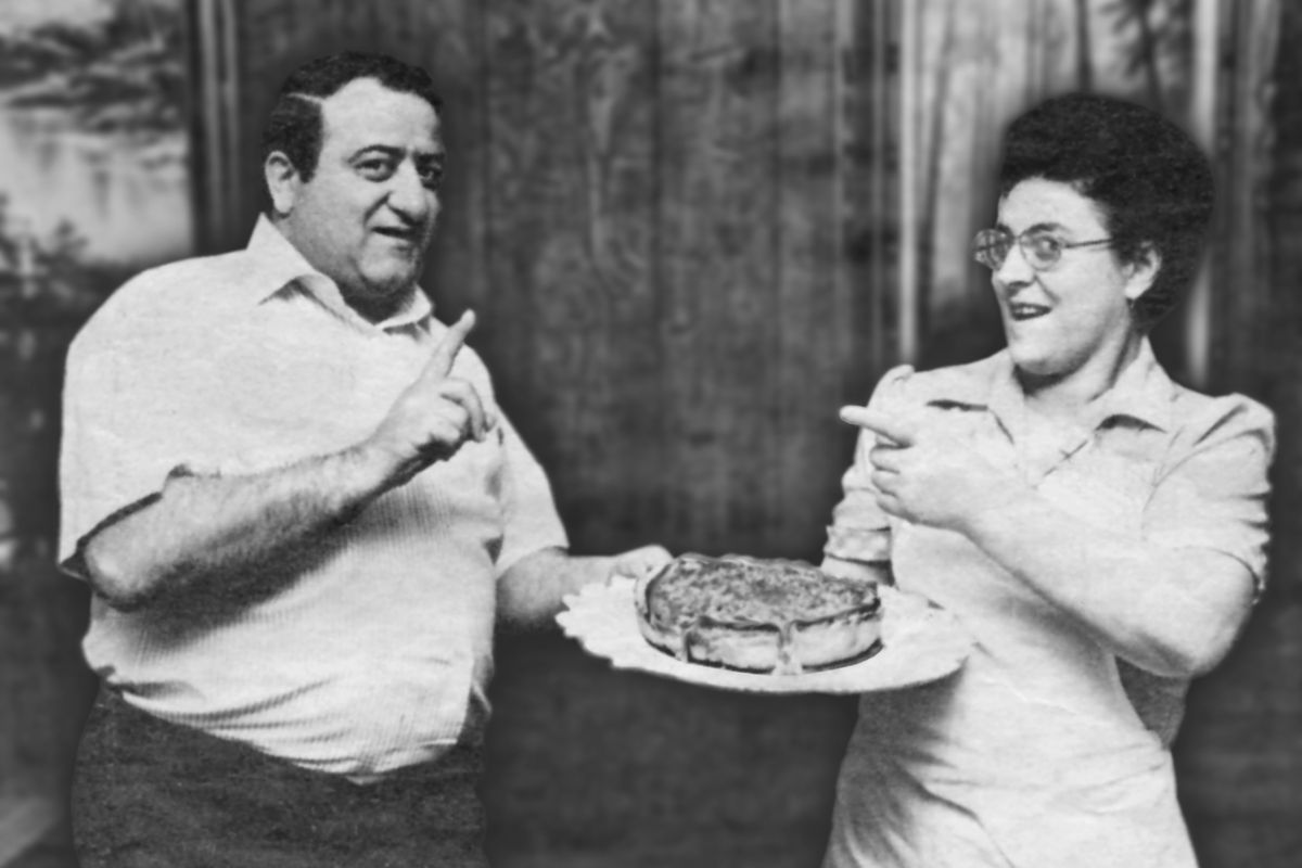 A man and a woman carry a plate containing a stuffed pizza and point at each other