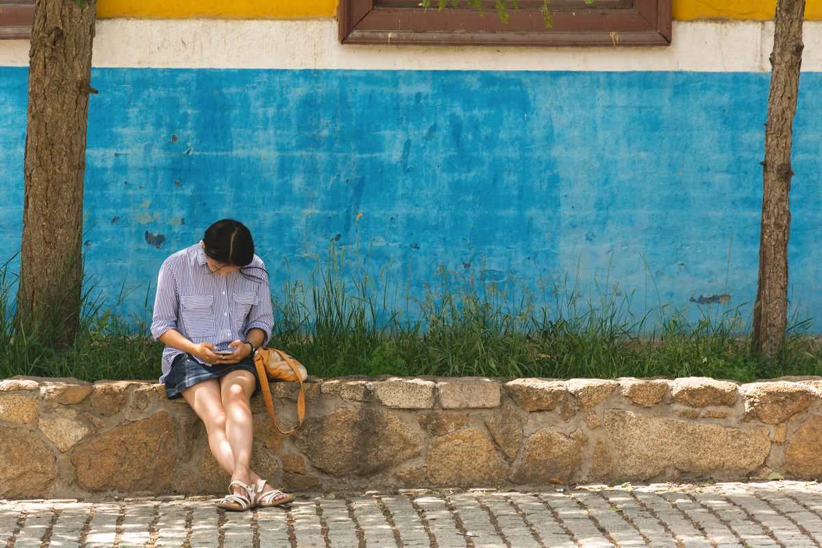 A person sits on a low wall looking at their phone.