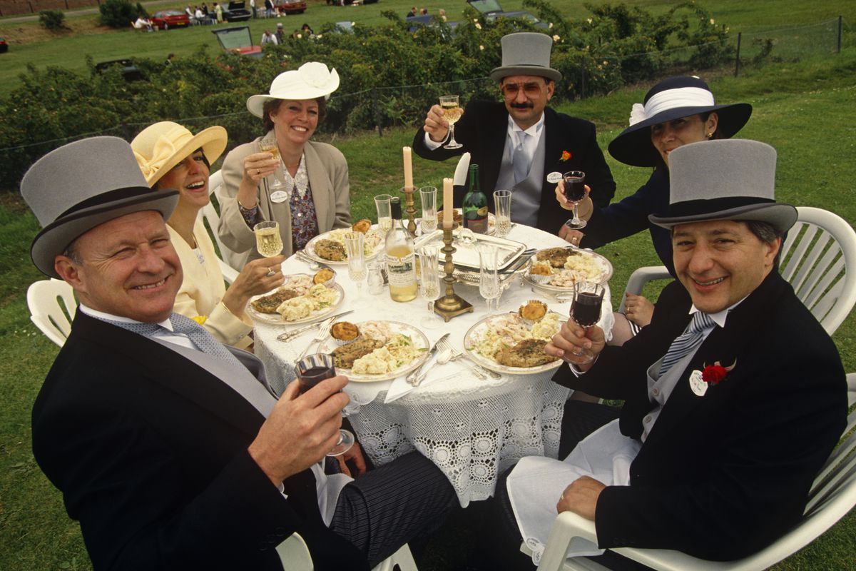 Three fancily dressed English couples sit around a white-clothed round table, at a formal picnic lunch for Royal Ascot racegoers on Ladies’ Day. 