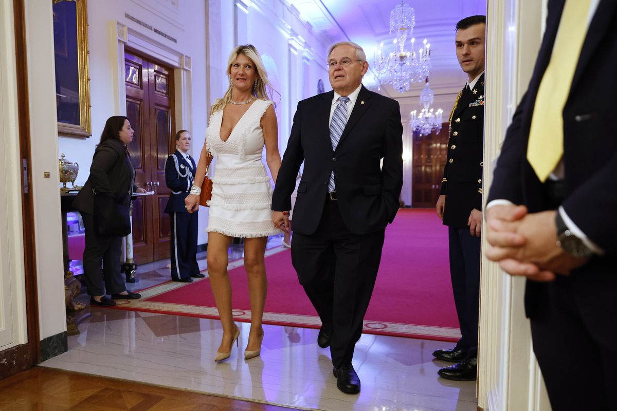 Senator Bob Menendez in a dark suit and his wife Nadine Arslanian in a white minidress and heels walk hand in hand down a White House hallway. 
