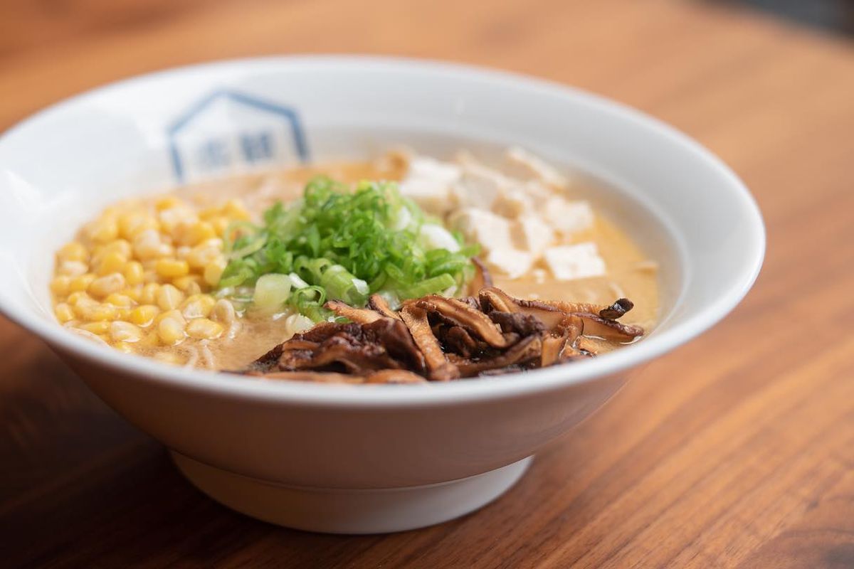 A white bowl of ramen garnished with tofu, green onions, corn, and sliced mushrooms. The bowl is on a wood table.