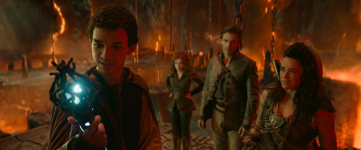 LtR: Justice Smith plays Simon, Sophia Lillis plays Doric, Michelle Rodriguez plays Holga, and Chris Pine plays Edgin in Dungeons & Dragons: Honor Among Thieves.  Simon raised his wand that glowed as the others looked on, in a cave full of rocks and lava. 