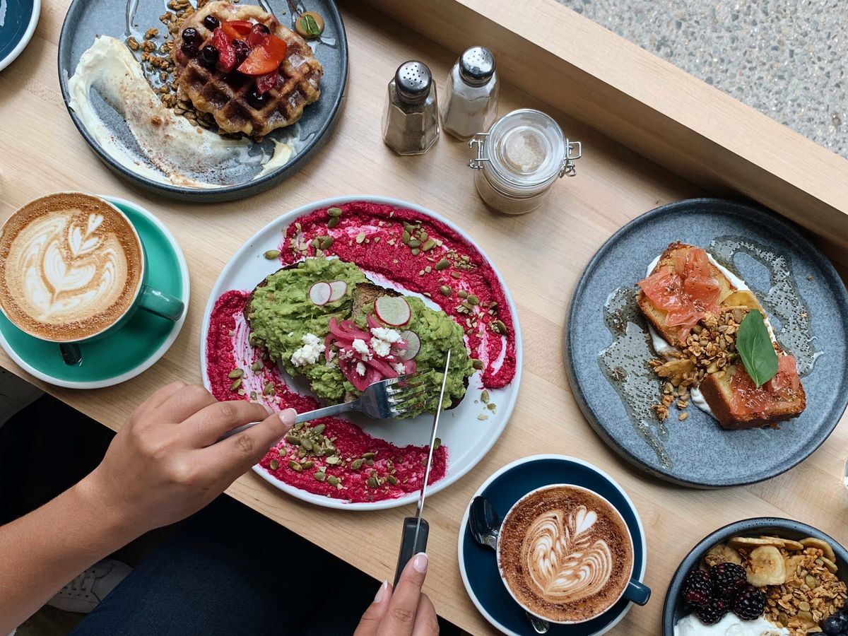 A person slices into an avocado toast plated with a beet puree, a side of waffles, a latte, brekkie bowls, and more.