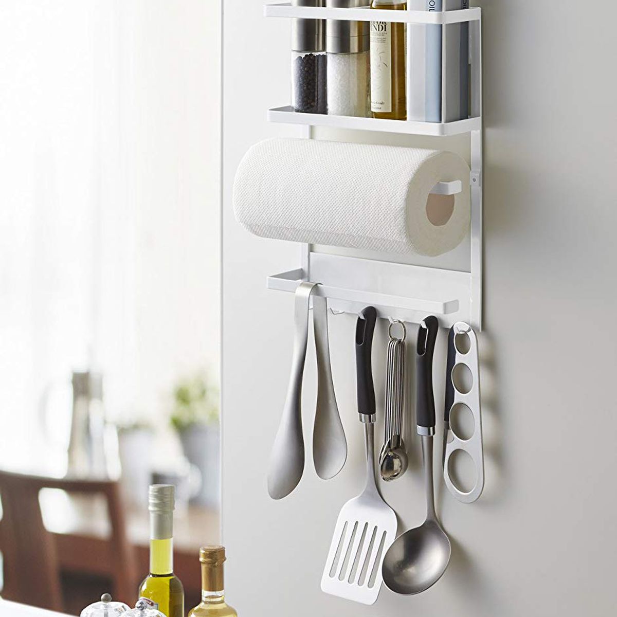 A white kitchen organization rack holds hanging cooking tools, paper towels, and salt and pepper. It’s magnetic and stuck on the side of a fridge.