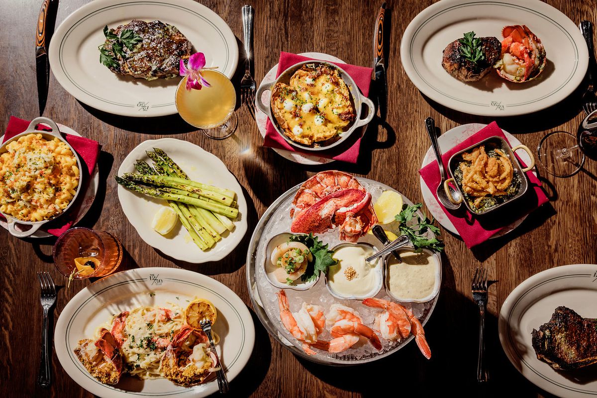 An overhead shot of a variety of plates on a wooden table featuring shrimp on ice, asparagus, steak, macaroni and cheese, and cocktails. 