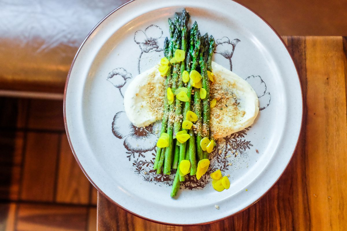 A plate of asparagus arranged over a creamy white sauce with some sort of crumble over the top and bright yellow sliced garlic. 