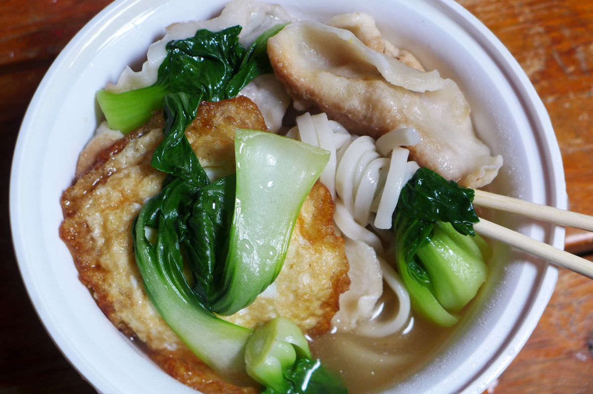 A soup in a white plastic bowl teeming with dumplings, noodles, and greens.