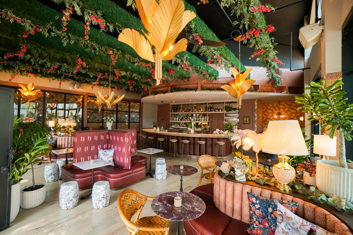 Inside Bar Eden, botanicals hang from the rafters, next to art deco lighting. The furniture is a mixture of marble side and bistro tables and pink booths of different materials and designs.