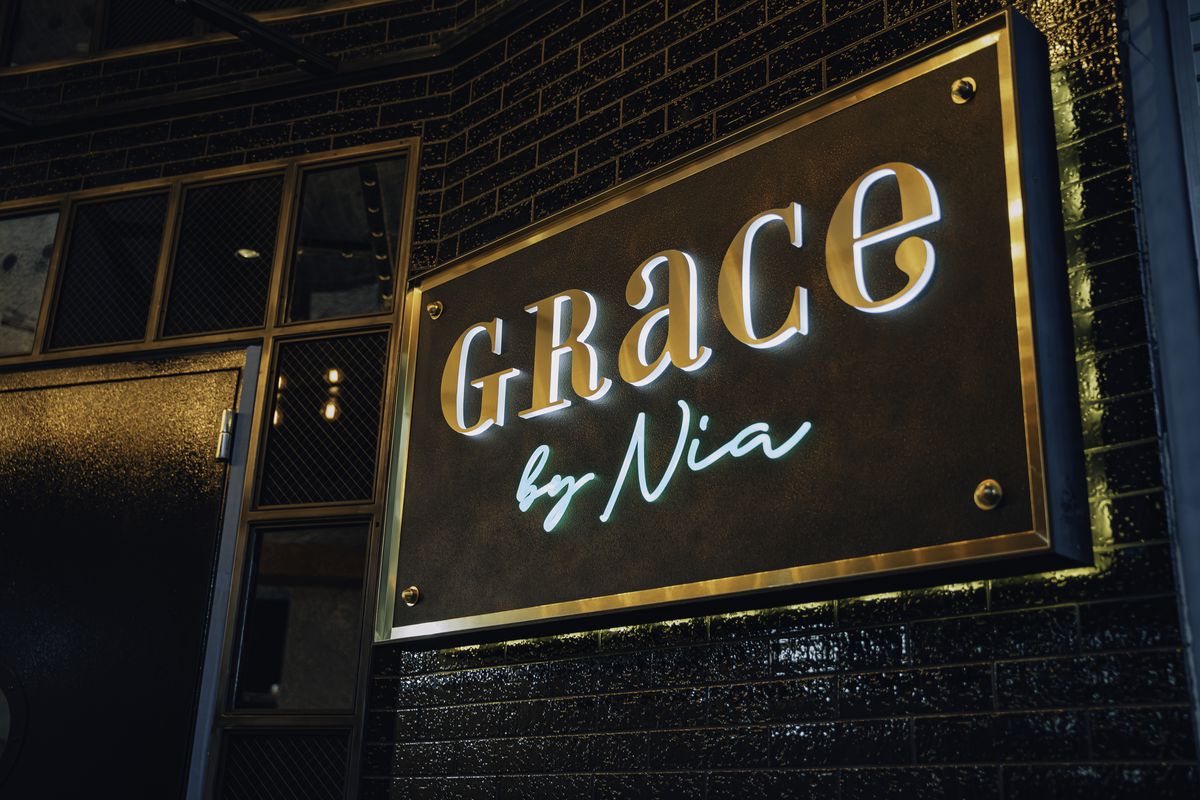 A gold and blue lit-up sign hanging on the black brick exterior of a restaurant that says “Grace by Nia”.