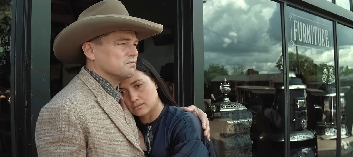Ernest (Leonardo DiCaprio, in tan suit and cowboy hat) comforts his wife Mollie (Lily Gladstone) as she leans against him in front of a furniture-store window in Killers of the Flower Moon