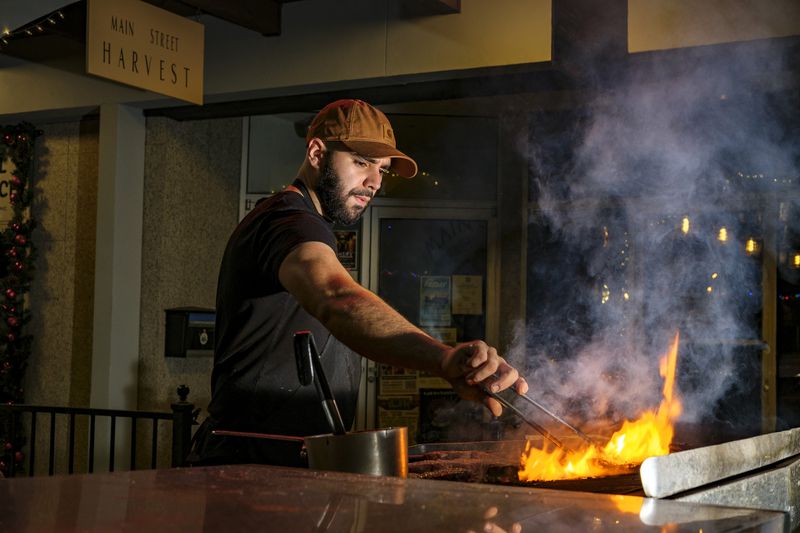 A man wearing a brown cap and black shirt sticks a pair of tongs into a large flame from a grill. 