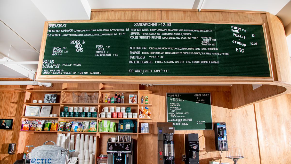 A coffee bar with blonde wood and a big peg board with sandwich menu items, then bags of chips, coffee mugs, and a percolator.