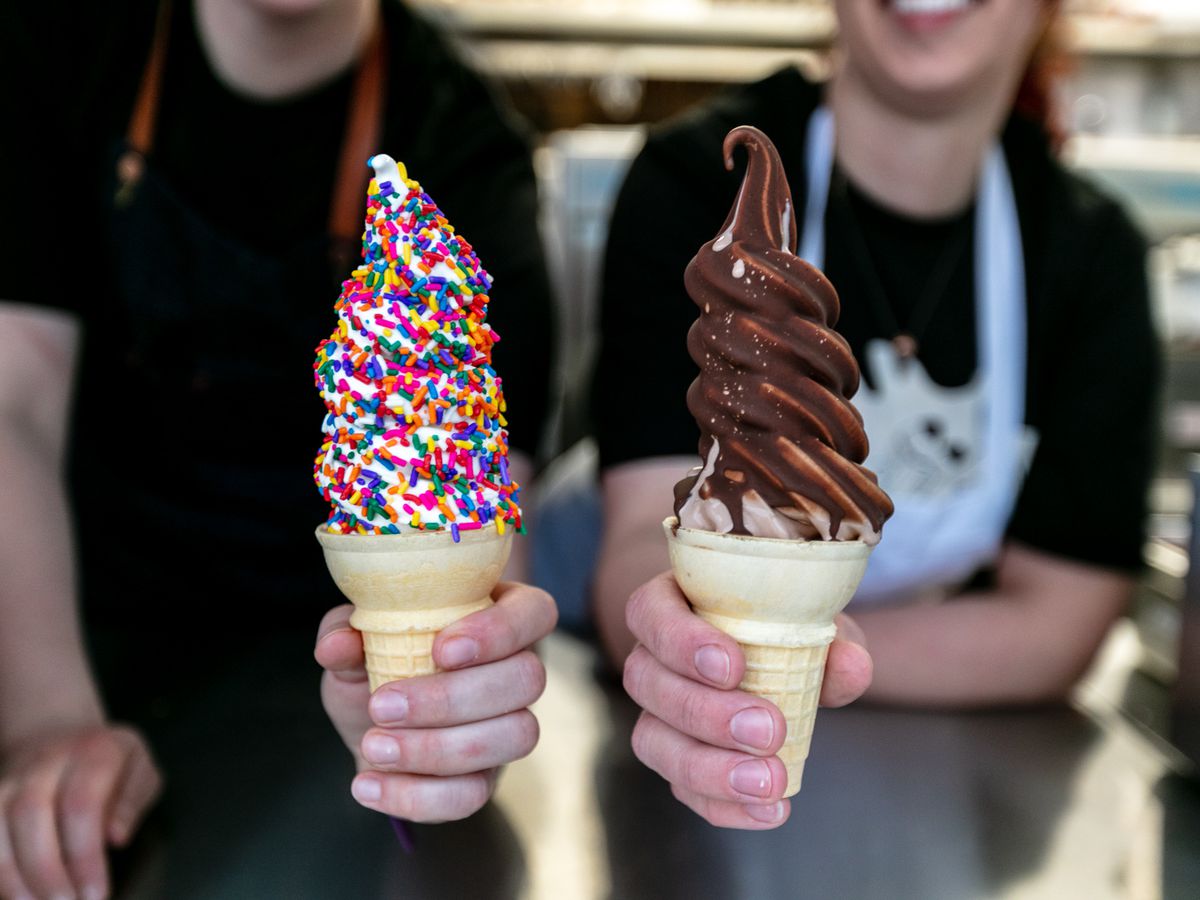 Two soft serve ice cream cones, one dipped in chocolate, one dipped in rainbow-colored sprinkles, held by hands from Doug’s Delight in Hazel Park, Michigan.