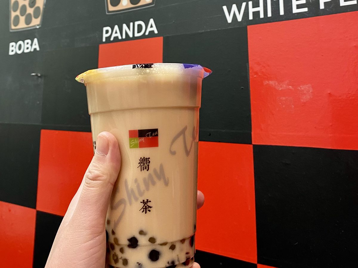 A brown bubble tea is held up on a red and black checkerboard backdrop.