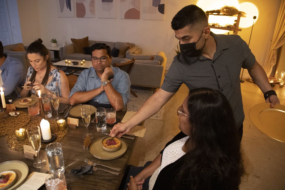 A man in a facemask places a bowl of soup in front of a woman who is sitting at the head of a rectangular table in a living room while other dinner guests look on