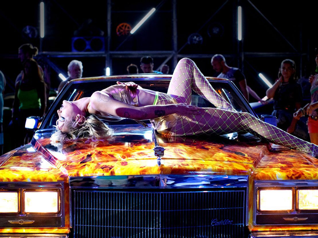 Una mujer joven se sienta a horcajadas sobre el capó de un automóvil pintado con luces de neón en Titane