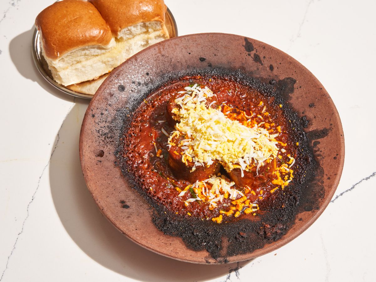 A wide-mouthed clay bowl of beef and red sauce with two golden buns on a small plate off to the side.