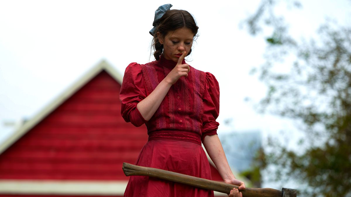 Mia Goth in a vivid red dress, holding an axe and doing a finger-to-the-lips “Shush” in Ti West’s Pearl