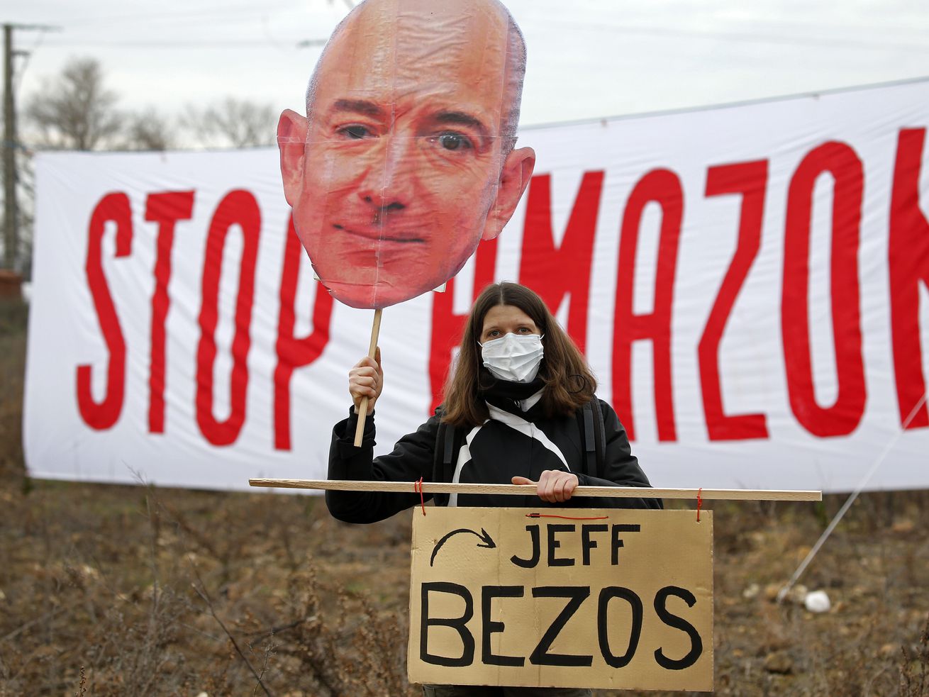 A protester holds up a large cutout of Jeff Bezos’s face while standing in front of a sign that reads “stop Amazon.”