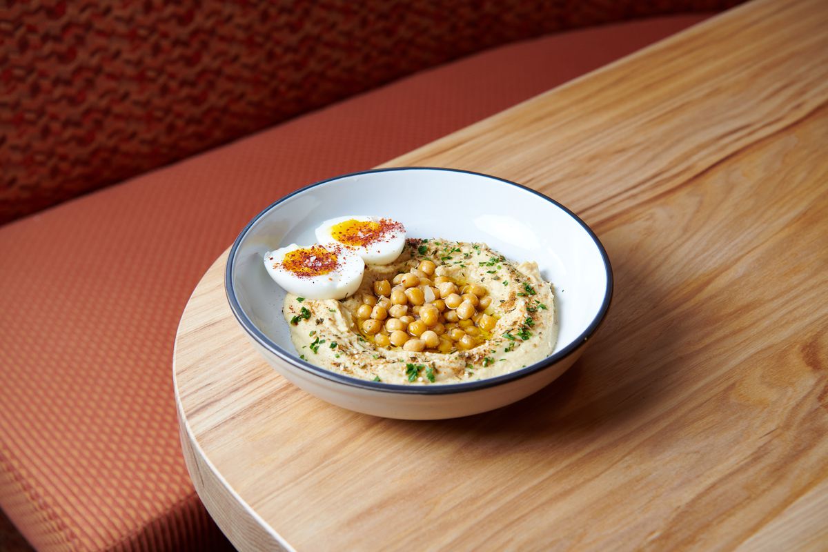 A bowl of hummus arrives garnished with olive oil, chickpeas, and a soft-boiled egg. 