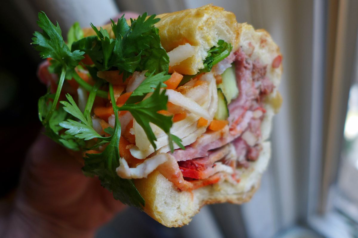 A bright banh mi sandwich seen in cross section with orange carrots, leafy deep green sprigs of cilantro, and layered meats.