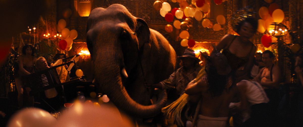 An elephant strides through a ballroom floor lit with dim red light, pushing through partygoers and balloons in Babylon