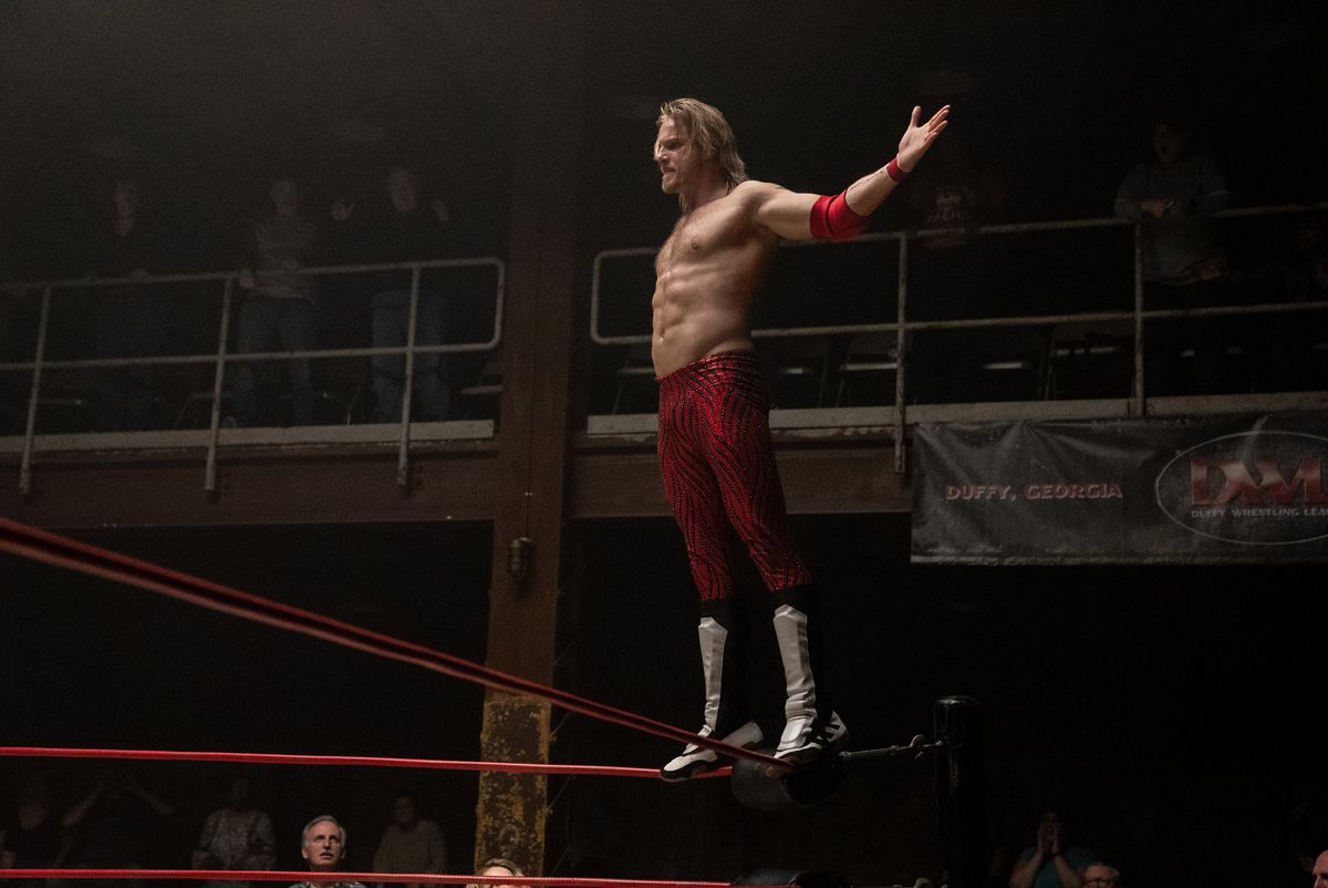 A wrestler stands on the ropes and holds his arms out for the crowd in a still from Heels