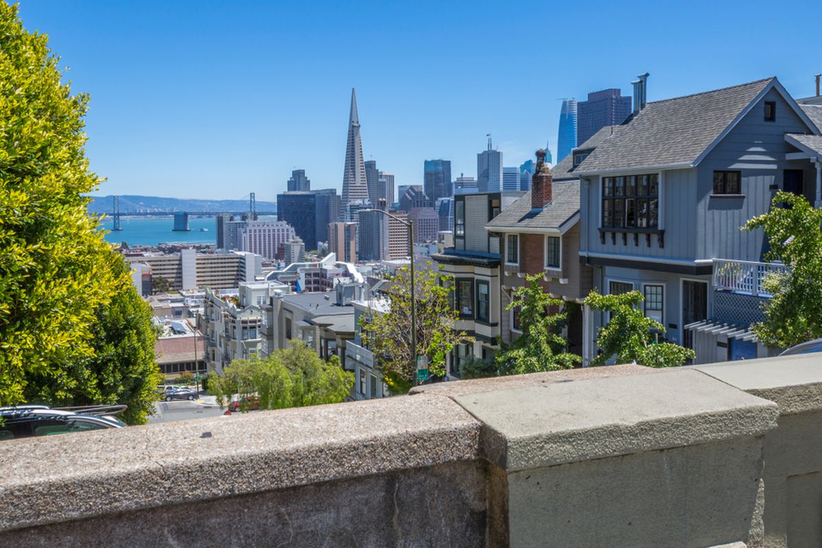 Homes and low-rise buildings in San Francisco, scene from the vantage point of a nearby ledge. Taller high-rise buildings—including one prominent one with a pointed peak—are visible further away.