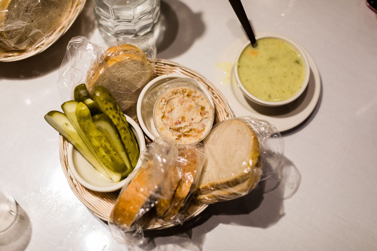 a plate with bacon spread, bread and pickles and a cup of yellow-colored dill pickle soup with a spoon in it
