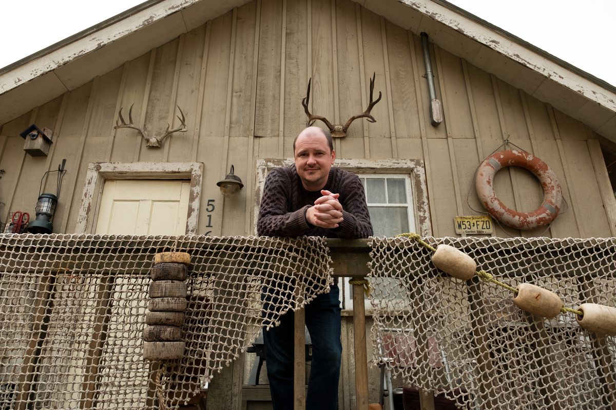 Mike Flanagan stands in front of a wooden house with antlers and a lifesaver on it. He leans against the railing.