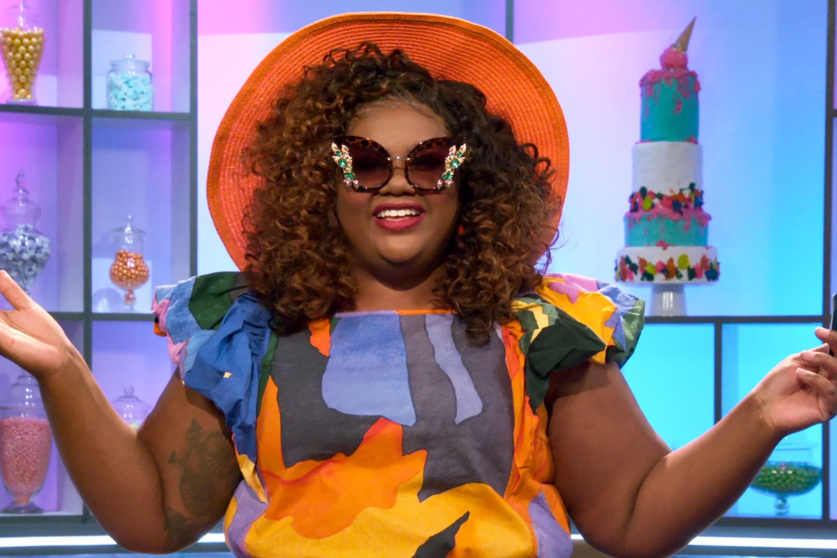 Nicole Byer, a woman wearing a bold floral print dress, fancy sunglasses, and a large orange hat 