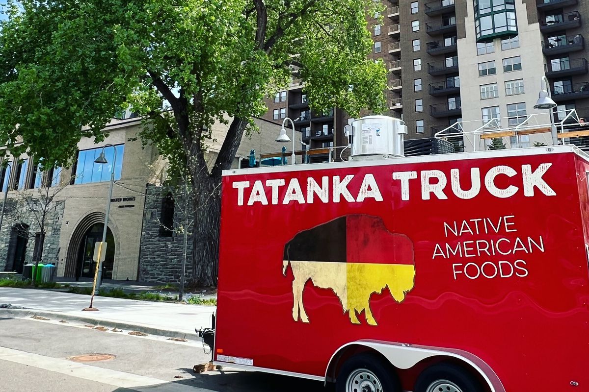The red Tatanka Truck, painted with a buffalo in the colors of the four directions (red, yellow, white, and black) and labeled with the words “Tatanka Truck Native American Foods” sits on a street on a sunny day. A tree and city buildings are visible in the background.