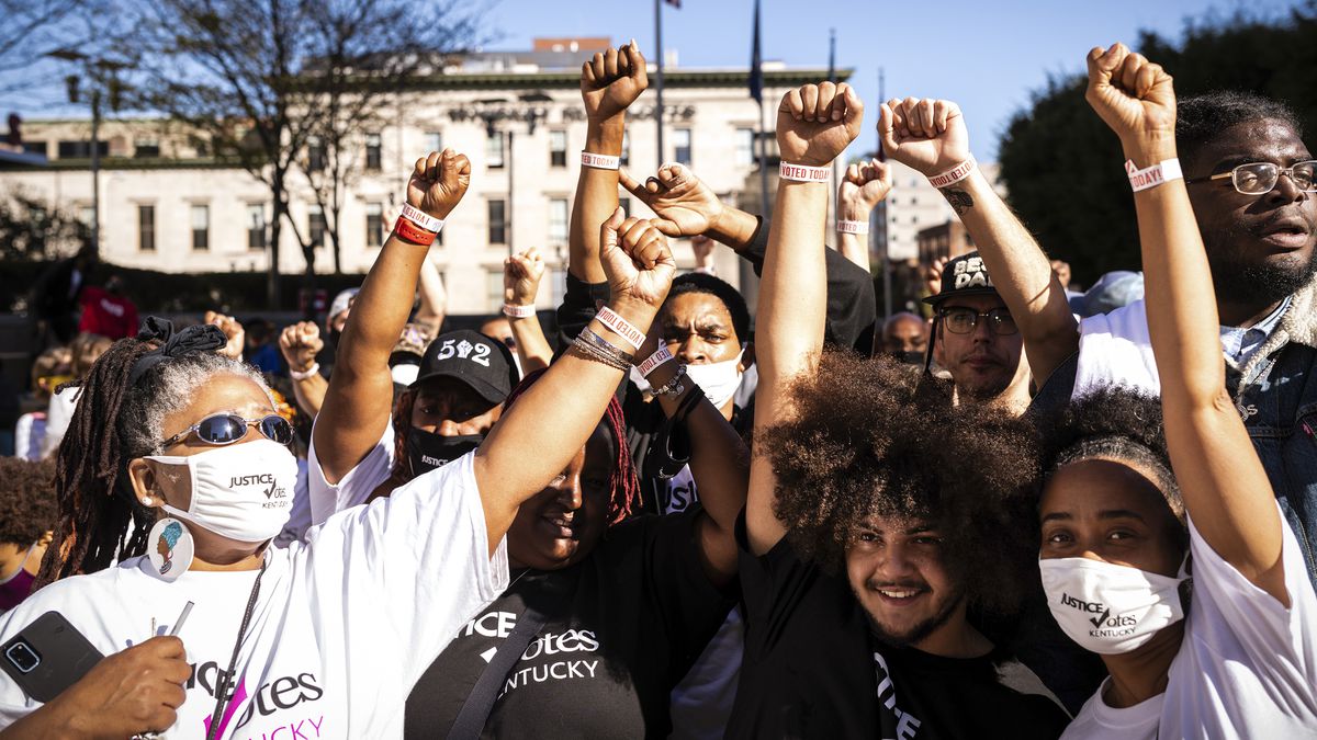 A crowd of about 14 Black people of various skin tones, all of them in either black or white, most of them wearing mask, and of varying age — some look as if they are in college; other have grey hair. All of them have one of their fists raised high, their wristbands visible in the day’s bright sun. Behind them is a flagpole flying the US flag.