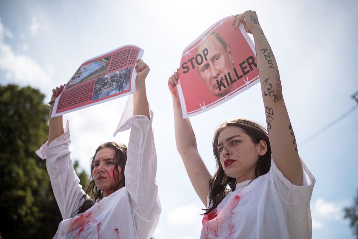 Two young women with fake blood on them hold signs over their heads. One is a picture of Vladimir Putin and the words “stop killer.”